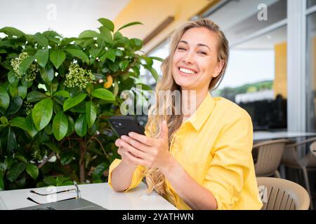 Happy freelancer woman holding smartphone while sitting at restaurant table. Beautiful middle age female professional is smiling and looking at camera during daytime outdoors.  Stock Photo