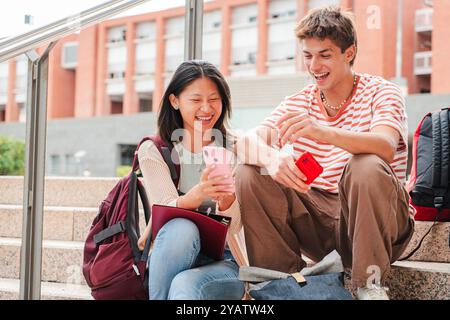 Two multiracial students sitting on a campus stairway, using a cellphone together. Casual outdoor scene of young friends watching a mobile phone while studying or relaxing in a university campus. High quality photo Stock Photo