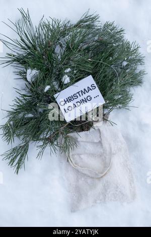 Paper note with text CHRISTMAS LOADING on Reusable shopping bag with fir spruce green branches in snowy outdoor winter forest background. Merry Christmas and new year concept. Natural bouquet greeting card copy space. Garland lights Stock Photo