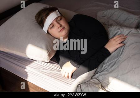 A 12-year-old boy is sick, he is lying on the bed under a blanket with a white wet cloth on his forehead. Stock Photo