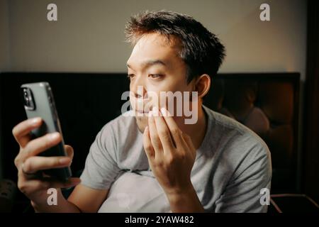 A man examining his facial hair with smartphone camera while touching his chin, appearing thoughtful and focused Stock Photo