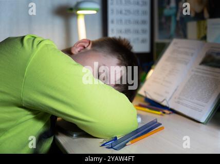 A boy in fell asleep while doing his homework. His head is lying on the desk, it is illuminated by a desk lamp, next to him is a pencil, pen, ruler. Stock Photo