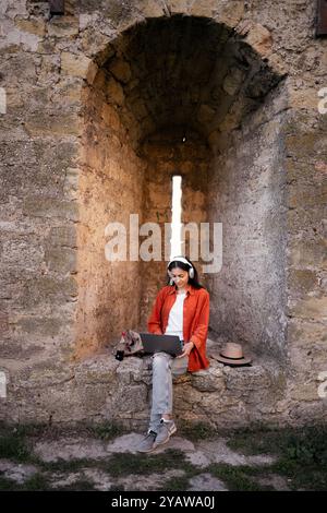 Traveler woman sits in ancient stone archway using laptop while wearing headphones. She enjoys remote work during her journey, combining technology wi Stock Photo