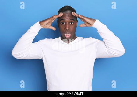 A Black man in a white sweater looks shocked, his hands on his head, eyes wide open, against a blue background, expressing strong surprise or disbelie Stock Photo