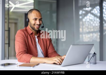 Smiling professional wearing headset works on laptop in modern office setting. Engaged in online meeting using phone technology. Notebook and pen on table indicate readiness and focus. Stock Photo