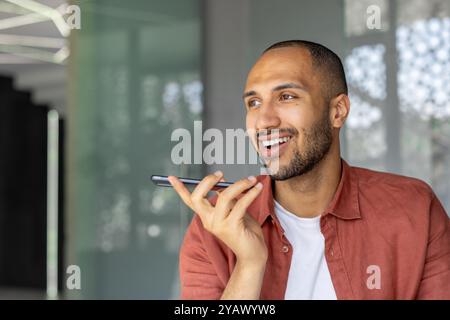 Smiling man using smartphone for voice call in modern office. Casual attire, expressing joy, connectivity. Professional, relaxed setting. Technology and communication theme. Stock Photo