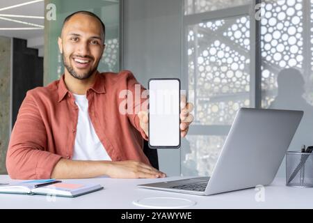 Smiling man holding smartphone displaying screen at desk with laptop and notebook. Shows use of technology and business in modern office environment. Ideal for communication concept. Stock Photo