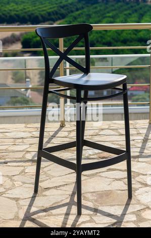 A stylish black bar stool stands alone on a sunlit terrace, with stunning views of verdant fields and trees in the background, inviting relaxation and Stock Photo