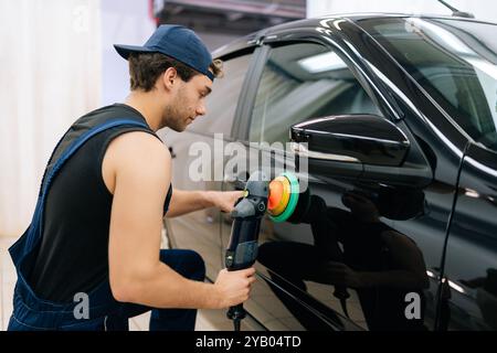 Rear view of muscular mechanic worker polishing door of luxury car in auto repair shop using with orbital polisher. Expert car paint technician using Stock Photo