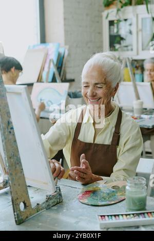 Vertical shot of silver haired woman smiling widely while doing strokes of paint brush on white canvas , she sitting at retro table with drawing tools Stock Photo