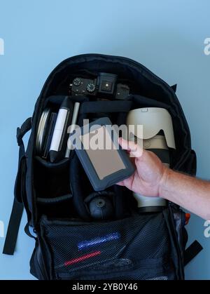A black camera backpack is open, revealing a DSLR camera, a lens, and other accessories. A male hand is holding an external hard drive. Stock Photo