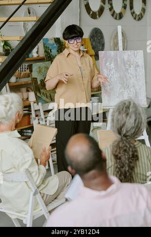 High angle shot of professional drawing teacher explaining new theme while senior students sitting on wooden chairs and taking notes Stock Photo