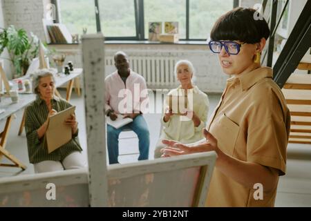 Young Caucasian teacher and senior biracial students looking at canvas while talented teacher explaining new topic Stock Photo