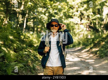 Senior male walking in the forest wearing eyeglasses and holding a hat. Mature man walking in the forest path. Stock Photo