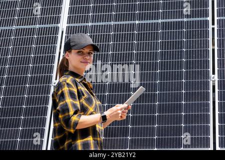 Female technician checking the operation of a solar panel, stands next to it and using digital tablet. Stock Photo