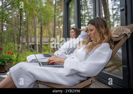 Two women enjoying remote work on laptops on a porch, embracing nature and cultivating a harmonious work-life balance at home. Radiating joy and tranquility Stock Photo