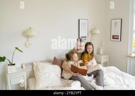 A father reads a book to his two daughters, who are seated on a bed in a comfortable bedroom. Stock Photo