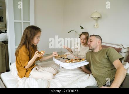 A father and his two daughters are enjoying a pizza meal together on a bed in their home. Stock Photo