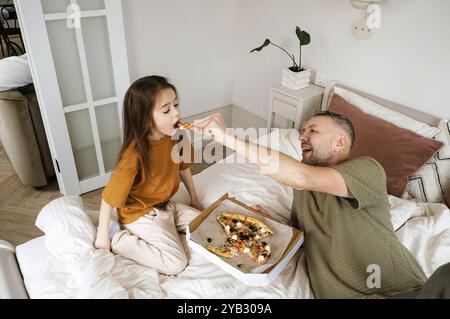 A father is sharing a pizza with his two daughters on a bed in their home. The father is happily feeding one of his daughters a slice, while the other Stock Photo