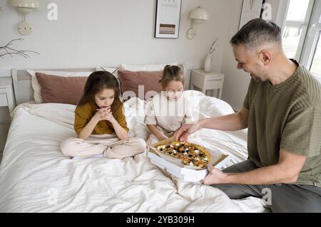 A father is serving his two daughters pizza in bed, creating a cozy and loving family scene. Stock Photo