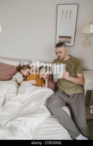 A father sits on a bed, reading a book on his tablet while his two daughters are sleeping peacefully. Stock Photo