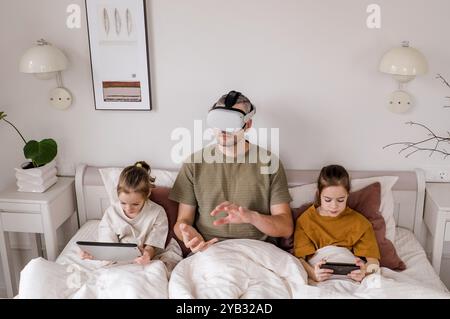 A father is experiencing virtual reality while his two daughters sit beside him, each engrossed in their own digital devices. Stock Photo