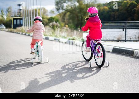 Two young girls ride bicycles with training wheels along a sunny paved path, surrounded by nature, wearing colorful jackets and helmets, illustrating Stock Photo