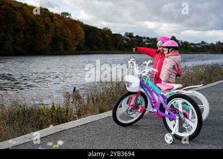 Two children on a lakeside pathway pause during their bike ride to admire the view. They wear colorful helmets and jackets, indicating a joyous outdoo Stock Photo