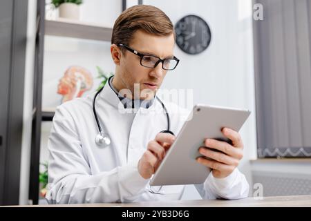 Male Caucasian doctor in white medical uniform filling patient medical history or anamnesis on tablet in office. Doctor consulting client online on ta Stock Photo