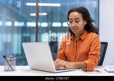 Focused woman in orange shirt working on laptop in modern office. Shows business, technology, and work concepts with phone and tablet on desk. Stock Photo