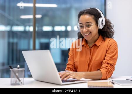 Female programmer wearing headphones, coding new software on laptop. Focused and happy while working in modern office. Ideal for technology, business concepts, inspiration. Stock Photo