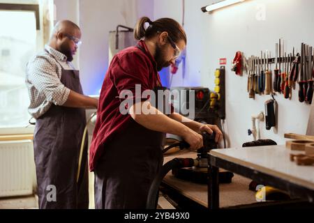 Carpenter and BIPOC coworker using angle grinder on lumber block to smooth surfaces by abrasion with sandpaper. Man uses orbital sander gear for furniture assembling, helped by colleague in background Stock Photo