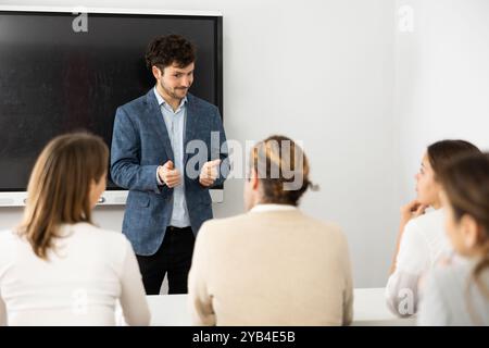 Male business coach standing near interactive board explaining topic while standing against group of people sitting at desks in front of him Stock Photo