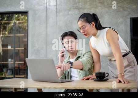 A handsome, serious Asian businessman is multitasking, talking on the phone with someone while reading on his laptop and discussing work with his coll Stock Photo