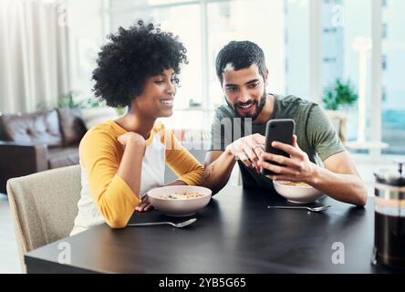 Eating breakfast, phone and happy couple in home for morning news, online shopping or social media. Food, man and interracial woman with mobile for Stock Photo