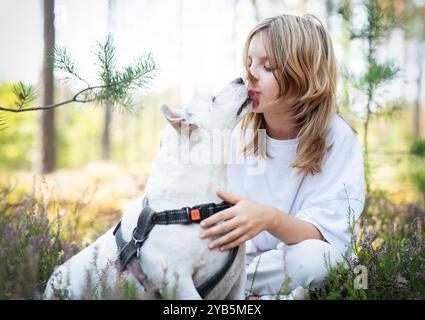 A young girl gently kisses her white dog while sitting among vibrant greenery. The serene atmosphere suggests a joyful afternoon bonding in the forest Stock Photo