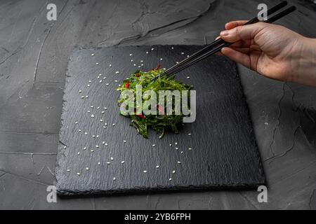 A woman's hand takes a seaweed salad with sesame seeds and chili peppers with chopsticks on a black mica board on a gray background. High quality phot Stock Photo