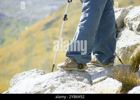 Outdoor activities: hiker standing on rocks at the edge of a mountain, close-up photo. Legs of a tourist who climbed to the top using trekking poles Stock Photo