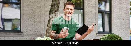 A young man sits comfortably outdoors, smiling as he writes in a notebook while using his phone. Stock Photo