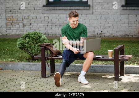 A young man with a prosthetic leg sits on a bench, smiling and focused while working on his laptop in the park. Stock Photo