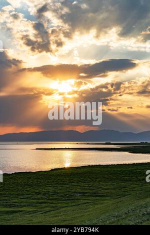 The sun's rays pass through the clouds and are reflected on the surface of the lake. There are mountains in the background. Stock Photo