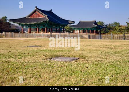 Gyeongju, South Korea - November 11th, 2023: An ancient pillar foundation is visible in the grassy foreground, with two traditional Korean pavilions s Stock Photo