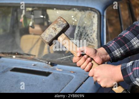 man stands with a hammer in his hands right in front of an old car with broken glass, ready to scrap it Stock Photo