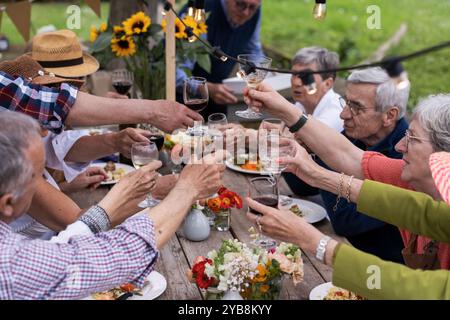 Group of lively senior friends toasting with wine glasses during an outdoor lunch party. Elderly people enjoying a meal in a garden setting with sunfl Stock Photo