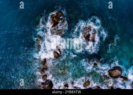Aerial view of powerful ocean waves crashing against jagged rocks in crystal-clear turquoise waters. The white foam contrasts beautifully with the dee Stock Photo