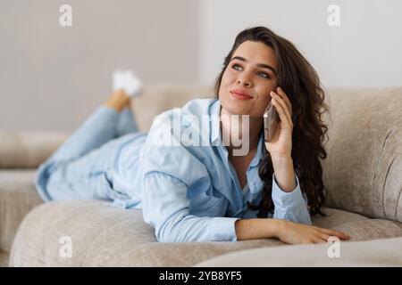 A relaxed young woman lies on a sofa, talking on a smartphone in a cozy, modern living room. Stock Photo