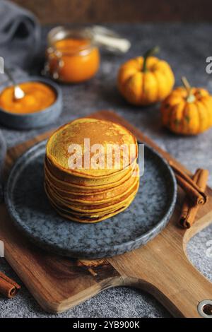 Light and fluffy pumpkin pancakes with honey and walnuts on a blue plate. Wooden cutting board, pumpkin pure in a glass jar, cinnamon sticks, and dark Stock Photo