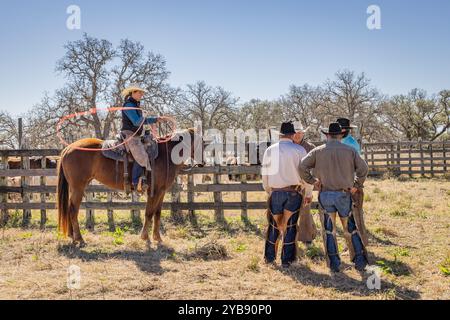 Yoakum, Texas, United States. March 15, 2022. Cowgirl on a horse swinging a lasso, with cowboys standing nearby. Stock Photo