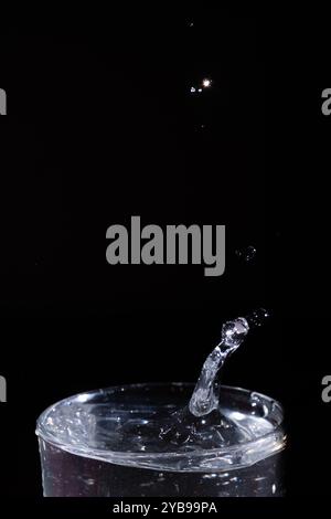 Drops of water leap from a transparent glass, creating dynamic shapes as they splash upwards against a striking black background, showcasing fluid mot Stock Photo