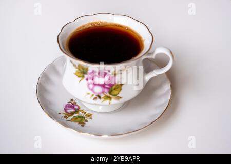 A beautifully crafted porcelain cup holds rich, dark coffee, resting on an elegant saucer adorned with colorful floral patterns, ideal for a serene mo Stock Photo